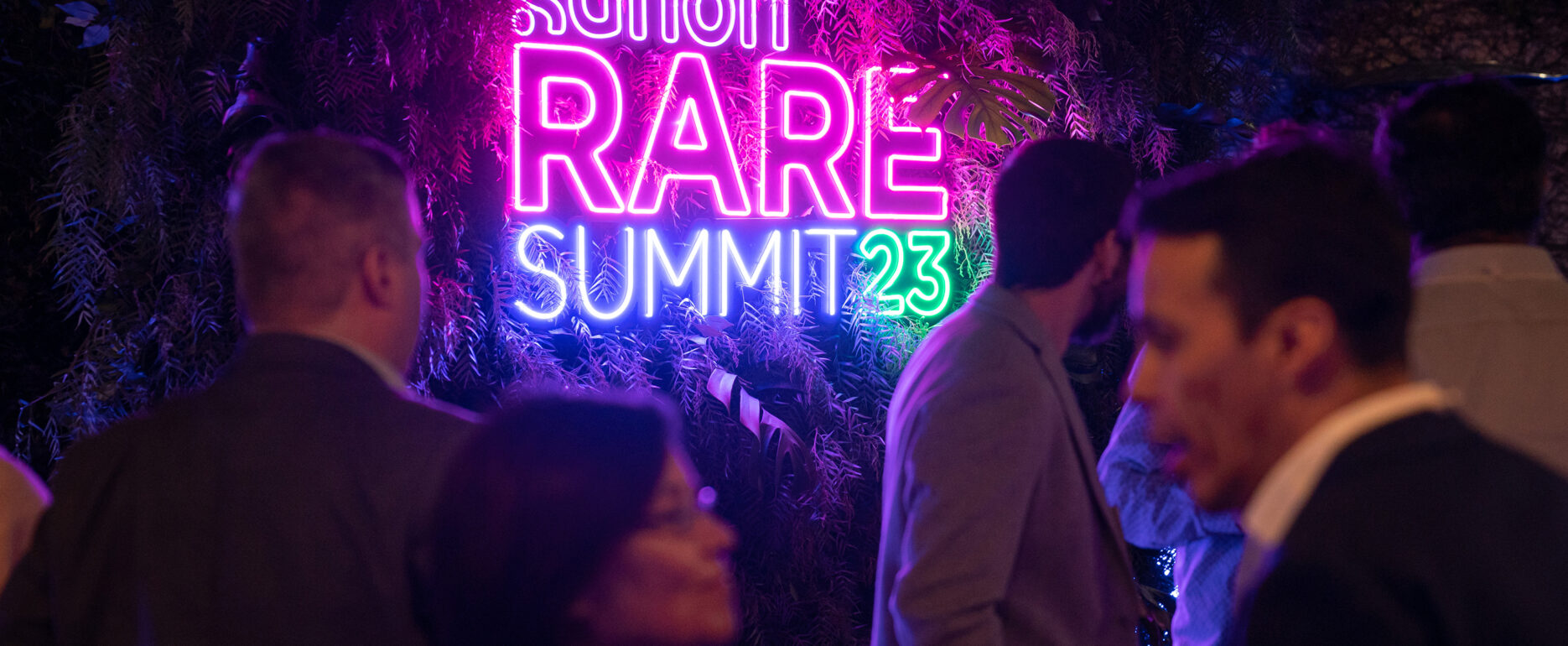 Un cartel neon que dice Sanofi RARE summit '23. Con gente hablando en segundo plano