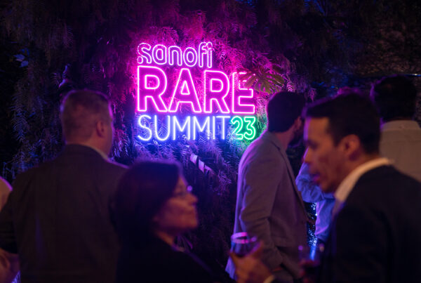 Un cartel neon que dice Sanofi RARE summit '23. Con gente hablando en segundo plano