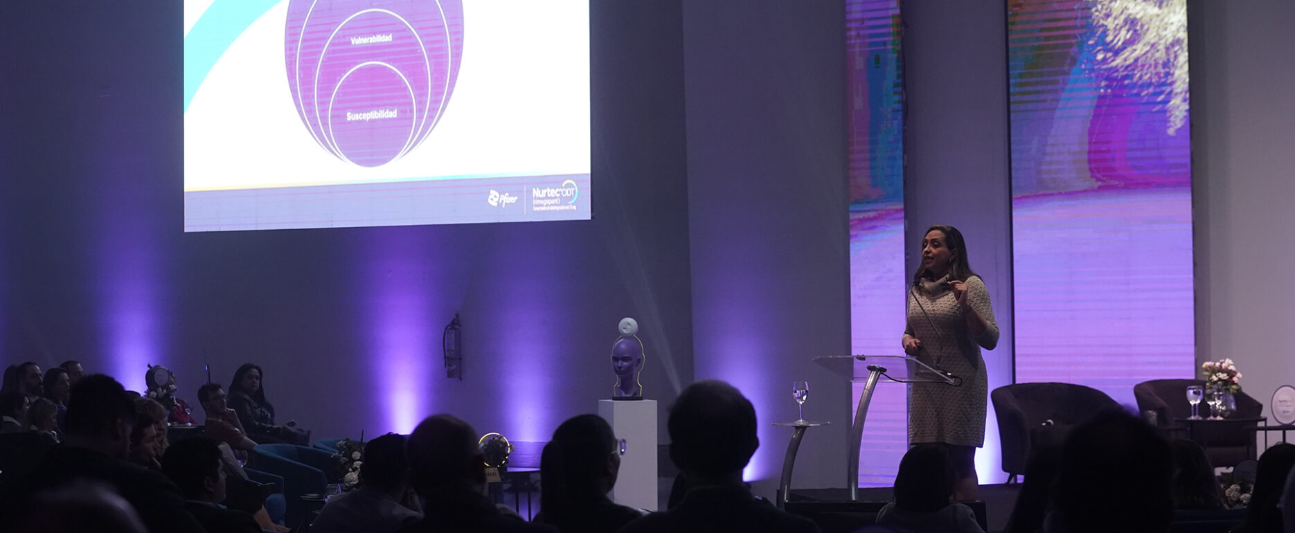 Persona exponiendo en un escenario con grandes pantallas mostrando una ppt