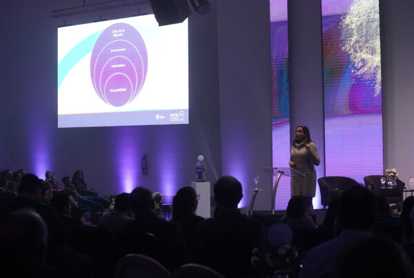 Persona exponiendo en un escenario con grandes pantallas mostrando una ppt